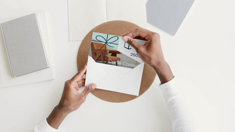 Man's hands at a desk putting a Qubik Furniture gift card worth a 250$ value into a white envelope.